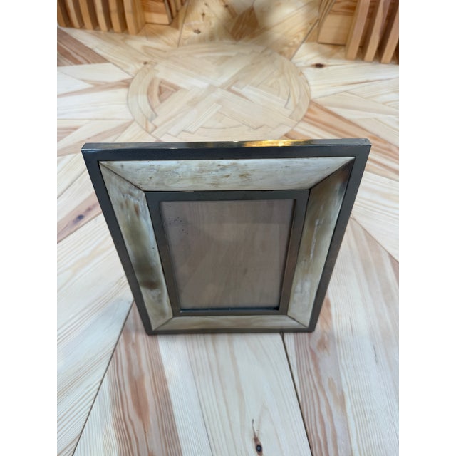 A vintage tabletop picture frame with a wide, mitered horn surround set between slim metal borders. The warm cream and...