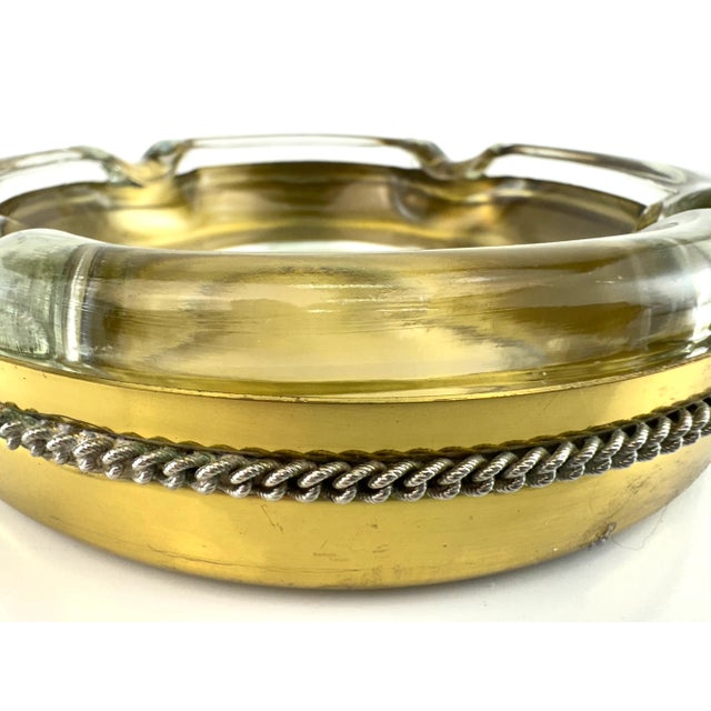 This striking mid-century ashtray features a thick, molded clear glass insert resting atop a polished brass base. The rich...