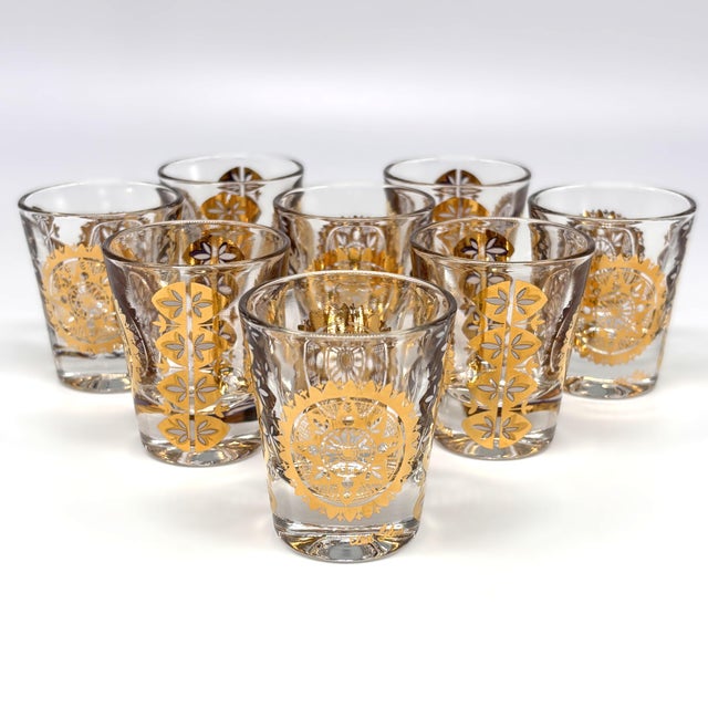 Set of eight vintage UTD shot glasses featuring bold gold medallion and stylized botanical motifs over clear glass, circa...