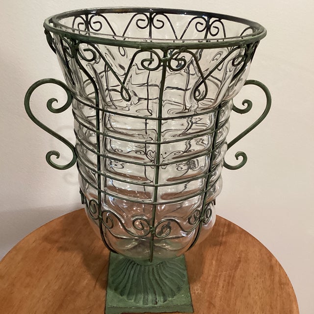 This large hand-blown caged glass footed vase features a wrought iron frame with scroll handles, making it a decorative...