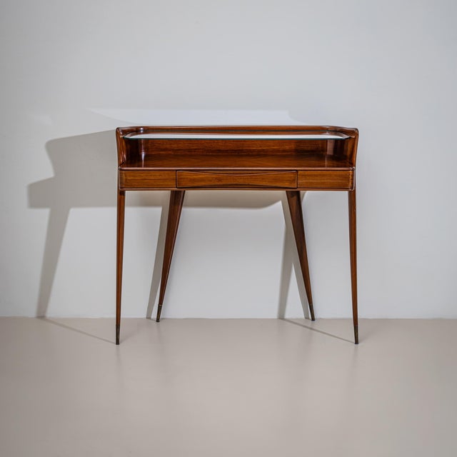 Elegant wall console raised on tall, tapered legs with brass sabots, featuring one drawer and a clear glass shelf. The...