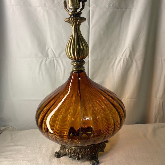 This is a vintage Hollywood Regency ribbed amber glass table lamp featuring an ornate cast brass footed base and neck.