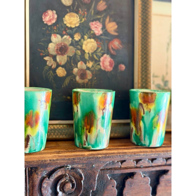 Striking set of six small ceramic cups featuring an expressive hand-glazed finish in rich green, amber, cream, and earthy...