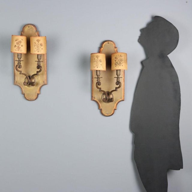 Pair of bronze and sheet metal wall sconces. Two lights supported by curved arms held up by a wooden panel covered in...