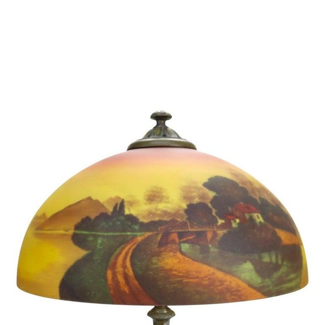 This antique table lamp features a reverse-painted glass shade depicting a scenic landscape, characteristic of the early...