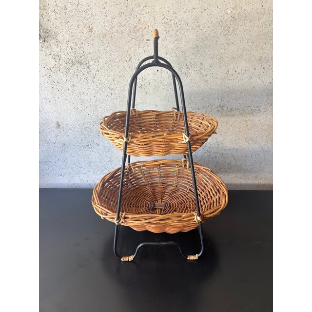 Vintage mid-century two-tier basket featuring handwoven rattan and cane baskets supported by a sculptural black iron...