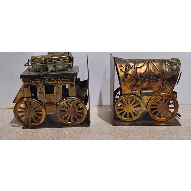 A vintage 1960s pair of copper-tone metal bookends with a Western theme: one depicts a Wells Fargo stagecoach with luggage...