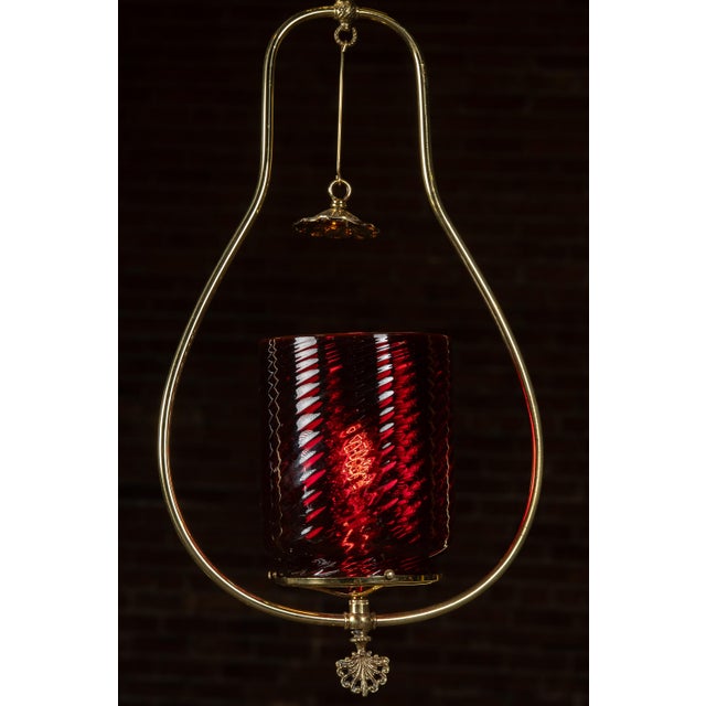 Pendant lamp with a sculptural brass frame in a teardrop outline, housing a ruby red glass shade with diagonal ribbed...