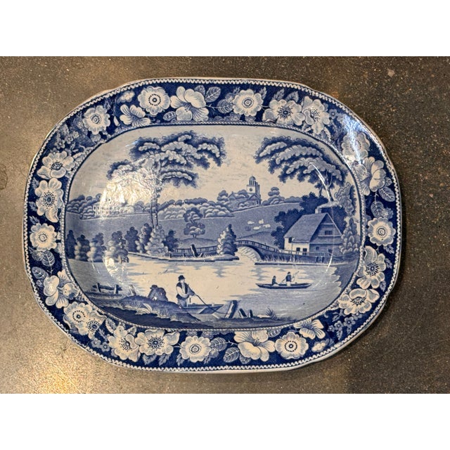 Blue transferware plate featuring a pastoral river landscape with a central boatman scene, framed by a dense floral...