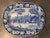 Blue transferware plate featuring a pastoral river landscape with a central boatman scene, framed by a dense floral...
