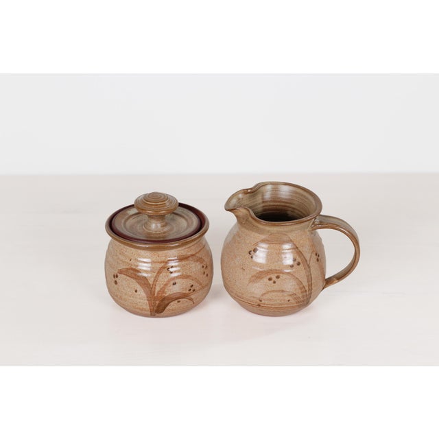 A 1988 vintage hand-thrown red stoneware creamer and lidded sugar bowl set, featuring a tan glaze with brown wheat motifs....