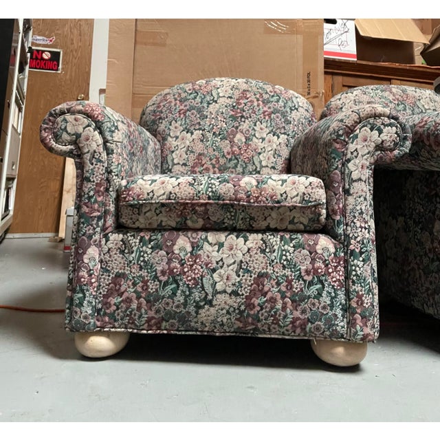 Large overstuffed arm chairs covered in a tapestry floral pattern featuring a dense mix of pink, mauve, cream, and green...