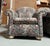 Large overstuffed arm chairs covered in a tapestry floral pattern featuring a dense mix of pink, mauve, cream, and green...