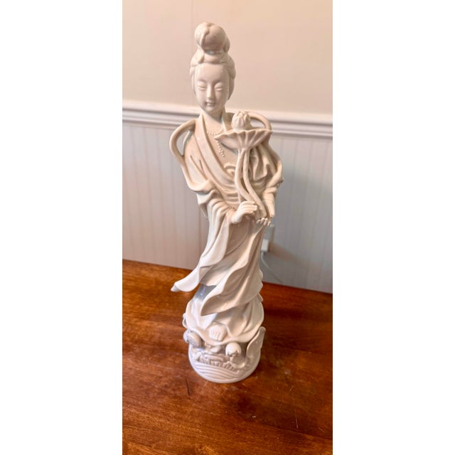 This statue of Guan Yin holding a lotus flower and standing in an elaborately carved base of lotus leaves and blossoms is...