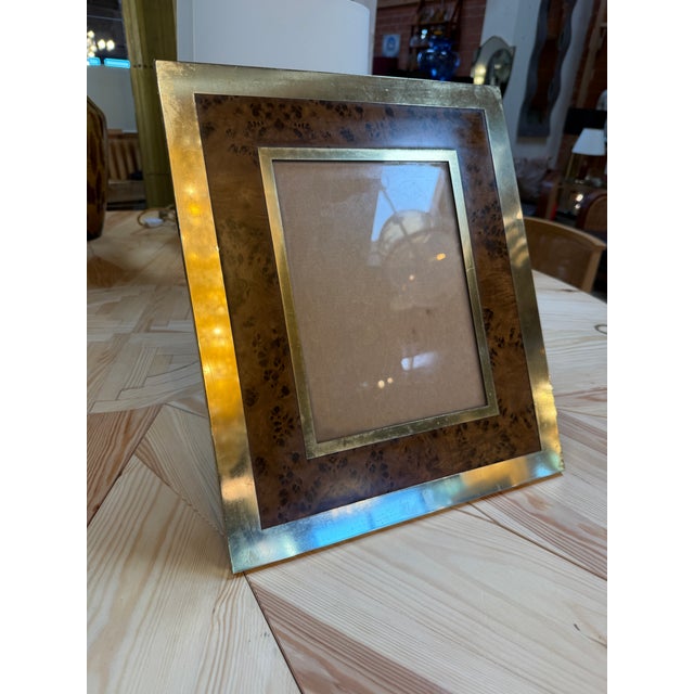 A tabletop picture frame with a richly patterned burl wood border, framed by warm brass trim on both the outer edge and...
