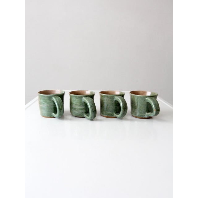 A set of four handmade vintage studio pottery mugs with a two-tone glaze in green and warm tan. Each cup carries subtle...