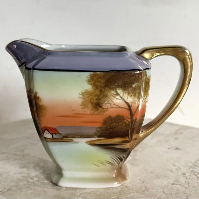 This Vintage Japanese Noritake Porcelain Creamer showcases a hand-painted pastoral scene, complete with soft sunset hues...
