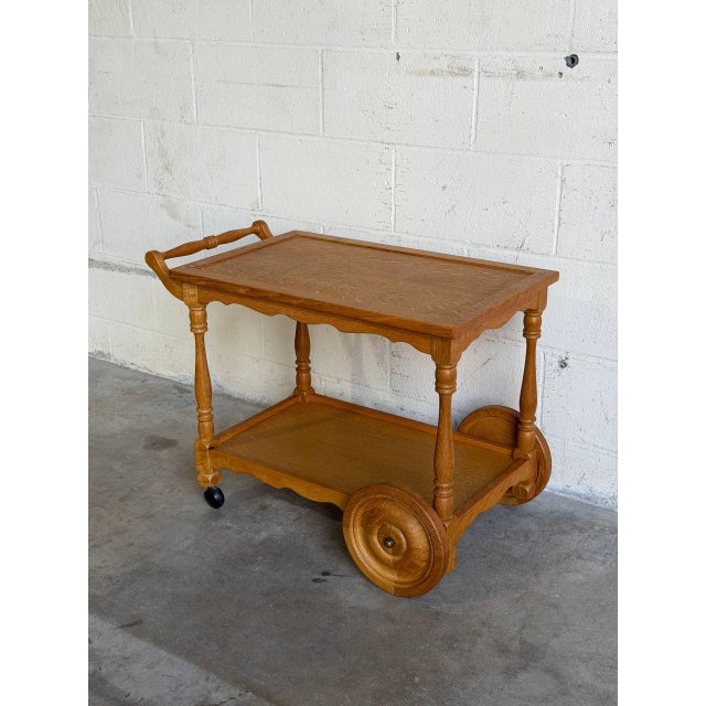 A mobile two-tier design features turned spindles, scalloped aprons, and large wooden rear wheels. An integrated push...