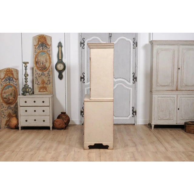A Swedish two-part tall cabinet from circa 1834 with off white/cream/light grey painted finish, two doors with carved...