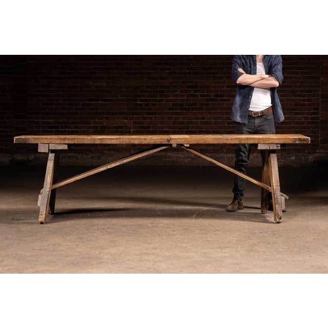 Long, low pine work bench constructed from thick, well-worn planks with a simple trestle base and pegged joinery. The top...