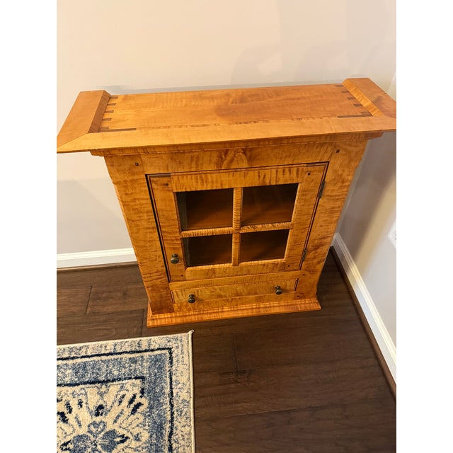 This piece is crafted from tiger maple wood and features a single door with a glass-front display.