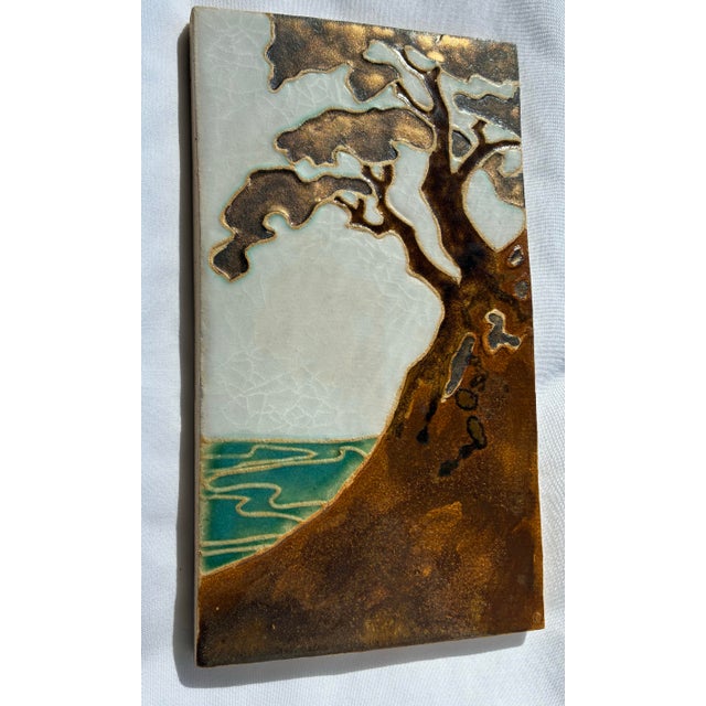 An exquisite Arts and Crafts style studio art pottery tile depicting a stylized cypress tree overlooking coastal waters....