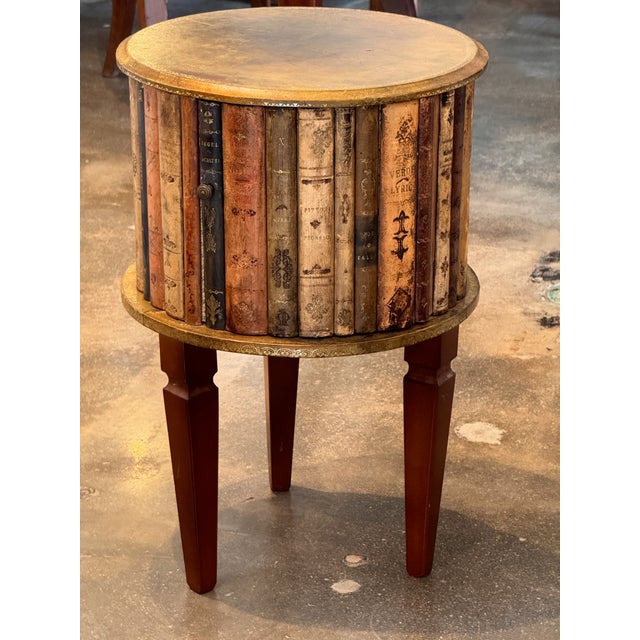 A round faux book table with a leather-covered top bordered by gilt decoration, above a cylindrical case formed as upright...