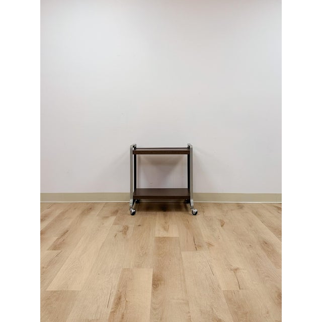 This mid-century bar cart combines a sleek chrome frame with dark wood laminate surfaces, capturing the clean, functional...