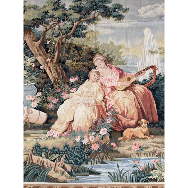 This large vintage tapestry presents an elaborate romantic garden scene with two elegantly dressed women surrounded by...
