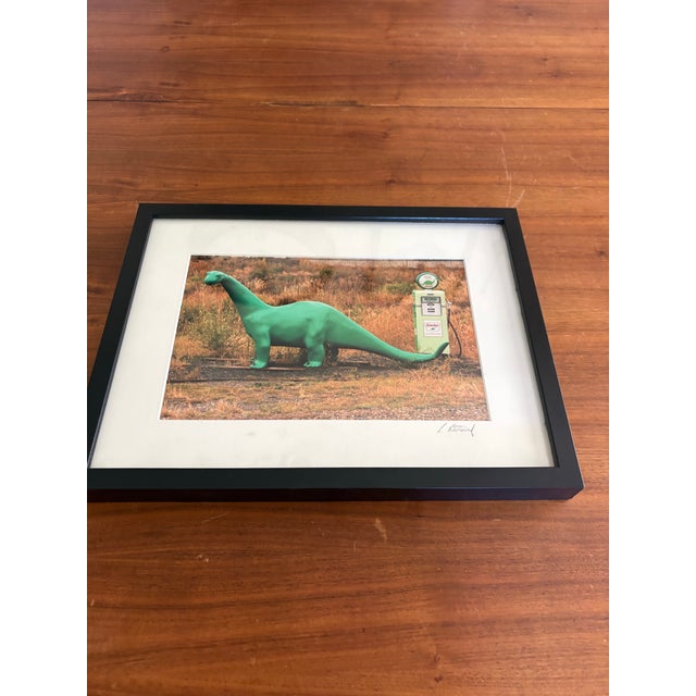 Whimsical and nostalgic framed color photograph featuring the iconic green Sinclair dinosaur posed beside a vintage “Dino”...