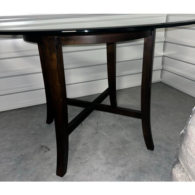 The table offers a circular glass top that is supported by a unique open-air wood base. Two crisscrossing stretchers...