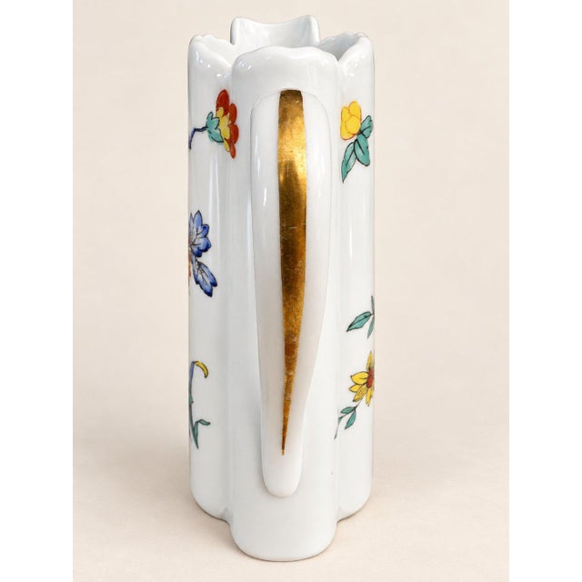 This is a white ceramic pitcher featuring a hand-painted floral design with red, yellow, blue, and green flowers and...