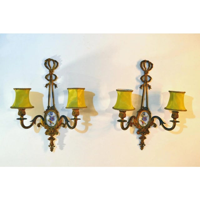 Gilded wall sconces in the shape of ribbons cast in solid bronze with inlaid hand-painted porcelain decor. Comes with lime...