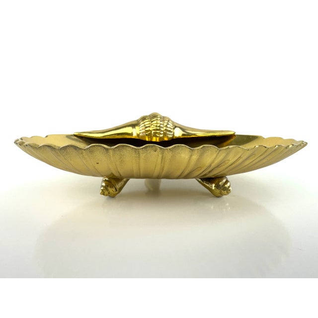 A charming vintage brass seashell dish featuring a raised conch shell detail and scalloped edge. Supported by three shell-...