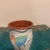 A hand-painted ceramic pencil holder featuring a whimsical, multicolored bird illustration against a cream-white...