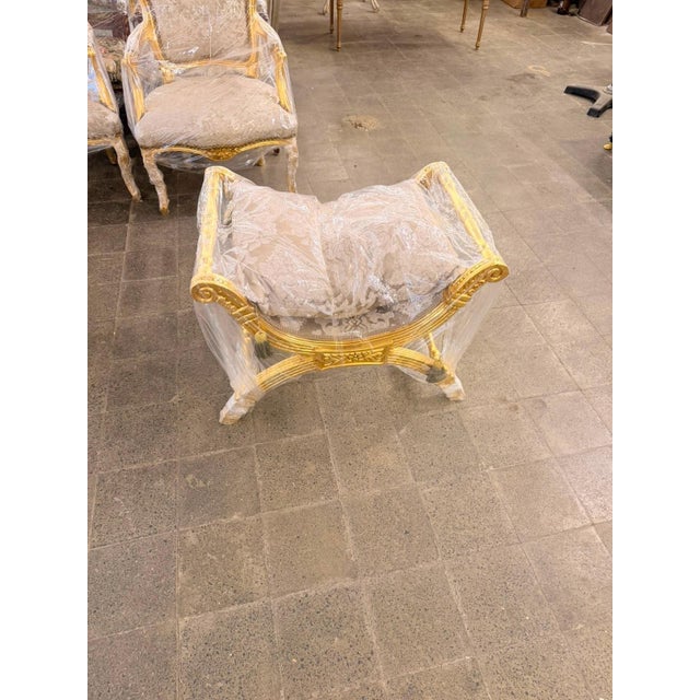 An elegant French Empire / Neoclassical curule-style bench, combining a sculptural gilded frame with soft blush-beige...