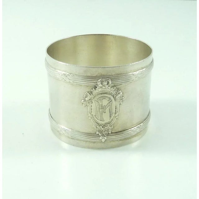 An elegant French silver napkin ring with cross ribbons over decorative bands. A large oval cartouche on the front is...