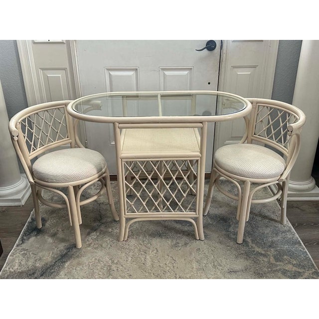 Charming vintage three-piece rattan bistro set featuring a sculptural glass-top table and two coordinating barrel-style...