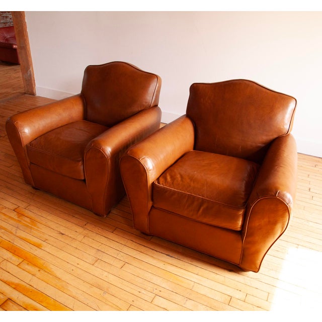 Fantastic pair of French Art Deco style club chairs from the 21st C. featuring beautiful brown-cognac leather and charming...