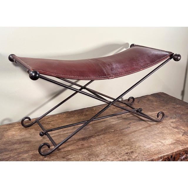 This vintage bench features a gently curved burgundy tinged brown leather seat stretched over a black wrought iron X-base...