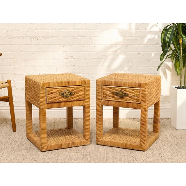 A charming pair of vintage rattan nightstands with woven surfaces, single drawers, and open lower shelves. Each table is...