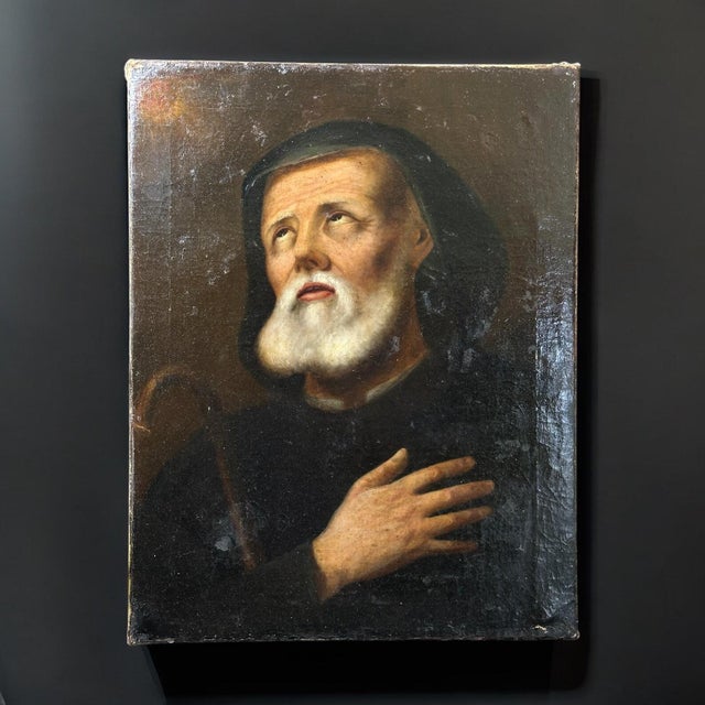 A solemn devotional oil painting depicting an elderly male saint, portrayed in a contemplative pose with his hand on his...