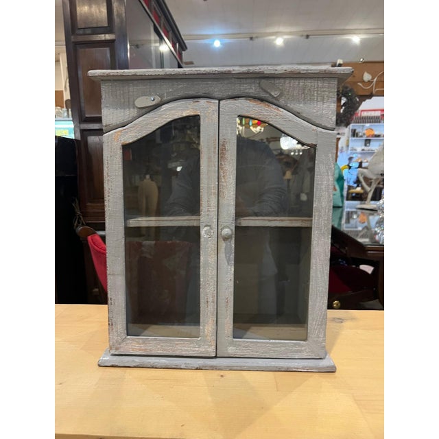 A charming vintage wooden wall cabinet with glass-paneled doors and a beautifully distressed painted finish. The weathered...