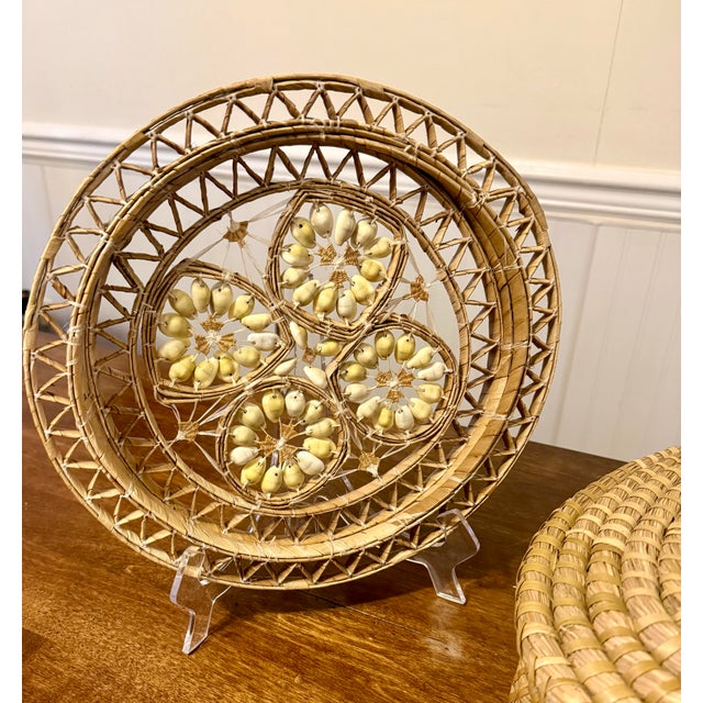 This is a circular, intricately woven decorative wall hanging adorned with numerous light-colored cowrie shells. Crafted...