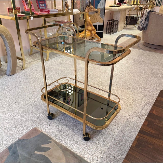 A beautiful vintage brass rolling bar cart in the style of D.I.A. The bottom shelf has mirrored smoked glass with a built...