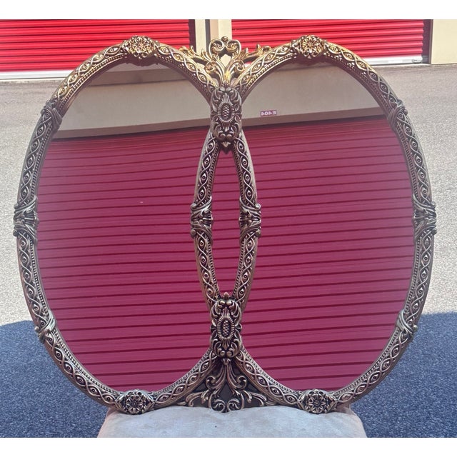 Striking and highly decorative, this Hollywood Regency-style wall mirror features an ornate double oval frame with...
