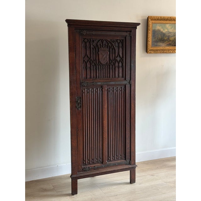 A tall Gothic Revival English oak corner cabinet, the full-height door carved with blind tracery around a crowned shield,...