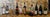 This oil painting presents a row of diverse bottles, each uniquely shaped and labeled, set against a textured background...