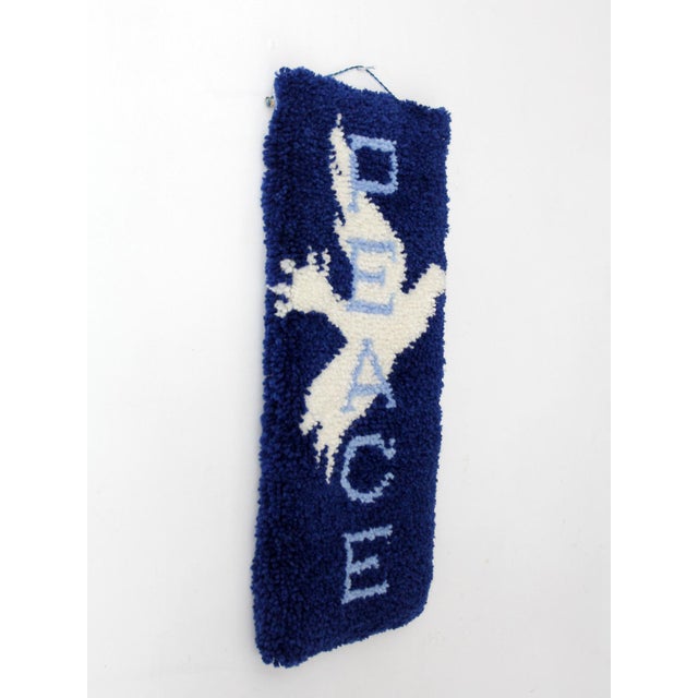 A vintage 1970s hand-hooked wall hanging featuring a soaring white dove and PEACE lettering on a rich royal blue ground....