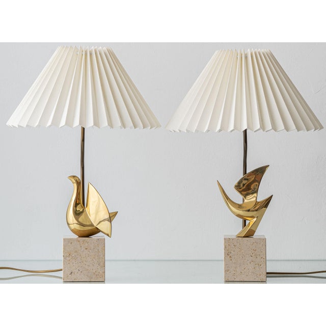 A kindred pair of French sculptural table lamps by Philippe Jean — polished brass bird forms on square travertine bases,...